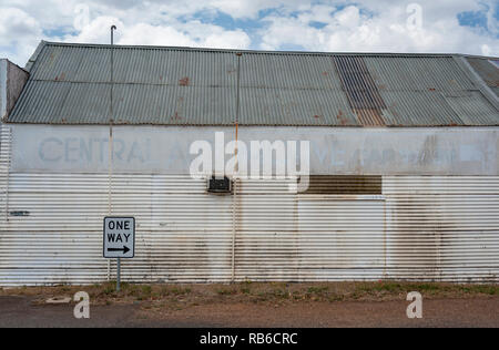 A One Way sign in from of an old corrugated walled building, silver and rust against a blue cloudy sky Stock Photo