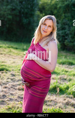 Pregnant woman in red dress standing outdoors and smiling Stock Photo