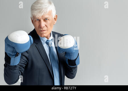 Senior business man wearing boxing gloves studio standing in defence position isolated on gray wall looking camera confident Stock Photo