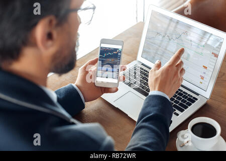 Mature man trader sitting at table at cafe with cup of coffee and laptop holding smartphone reading trading charts concentrated back view close-up Stock Photo