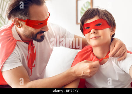 Father tying knot on little son cloak wearing superheroe costumes sitting together at home smiling dreamful close-up Stock Photo