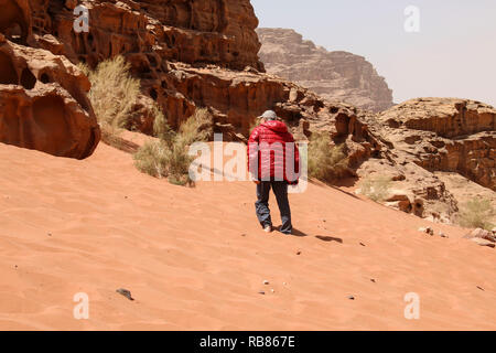 Man in a red jacket rises to a dune of red sand of the canyon of Wadi Rum desert in Jordan.