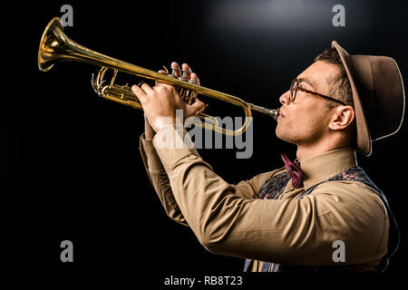 side view of young male jazzman in hat and eyeglasses playing on trumpet isolated on black Stock Photo