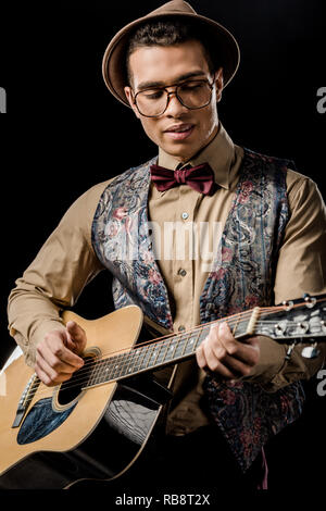 young male musician in eyeglasses and hat playing on acoustic guitar isolated on black Stock Photo