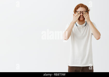 Studio shot of funyn good-looking tired male office worker rubbing eyes under glasses and smiling feeling exhausted after sitting near computer too long staying optimistic and joyful over gray wall Stock Photo