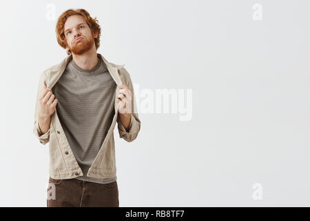Redhead guy picked wrong clothes feeling boiling hot in warm jacket breathing out waving with cloth, looking up at sun feeling discomfort standing displeased and bothered over gray background Stock Photo