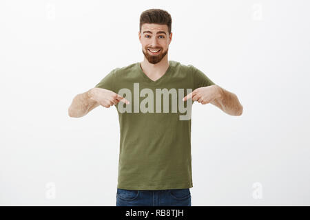 Portrait of cute adorable bearded boyfriend with charismatic smile grinning happily as pointing to centre looking at camera promoting copy space indicating at t-shirt over white background Stock Photo