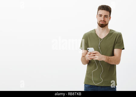 Man put on song brings nice memories. Portrait of charming cute and gentle bearded boyfriend in olive t-shirt smiling delighted with relaxed grin as listening music in earphones, holding smartphone Stock Photo