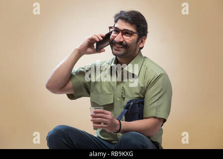 Smiling man wearing eyeglasses talking over mobile phone while holding a small pouch in one hand and a glass of tea in the other. Stock Photo