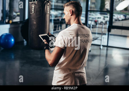 back view of personal trainer using tablet near punching bag in gym Stock Photo