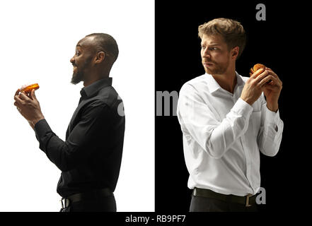 Men eating a hamburger and donut on a black and white background. The happy afro and caucasian men. The burger, fast, unhealthy food concept Stock Photo
