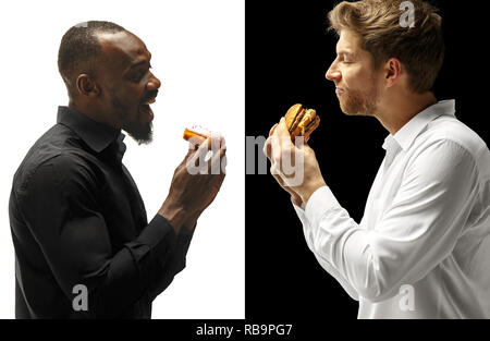 Men eating a hamburger and donut on a black and white background. The happy afro and caucasian men. The burger, fast, unhealthy food concept Stock Photo