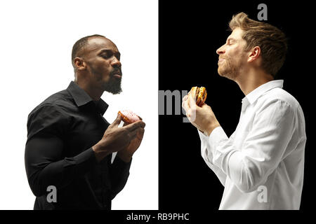 Men eating a hamburger and donut on a black and white background. The happy afro and caucasian men. The burger, fast, unhealthy food concept Stock Photo
