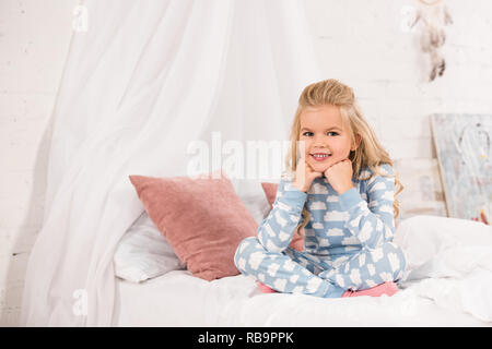 smiling cute kid sitting on bed with crossed legs and hands near face Stock Photo
