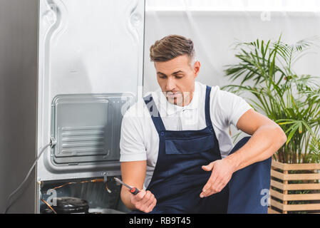 confused adult repairman looking at screwdriver while repairing refrigerator Stock Photo