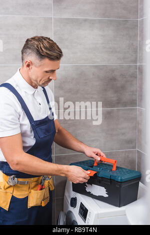 repairman in tool belt closing tools box after repairing washing machine at bathroom Stock Photo