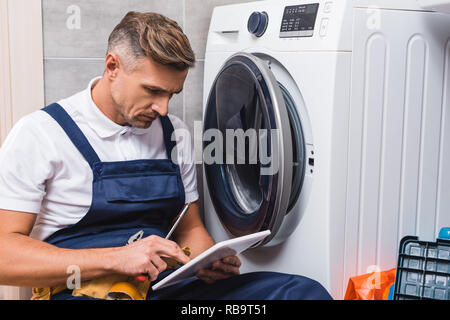 adult repairman holding screwdriver and using digital tablet while repairing washing machine in bathroom Stock Photo