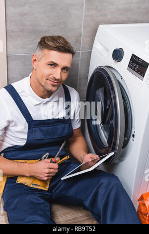 adult repairman holding screwdriver and using digital tablet while repairing washing machine in bathroom and looking at camera Stock Photo