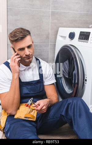 adult repairman holding screwdriver and talking on smartphone while repairing washing machine in bathroom Stock Photo