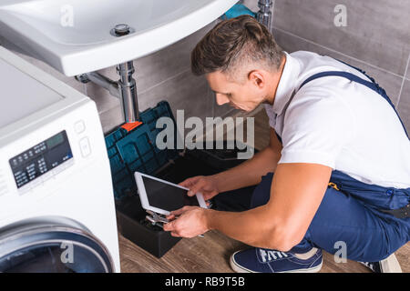 adult repairman sitting on floor and using digital tablet while repairing sink at bathroom Stock Photo