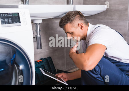 thoughtful adult repairman sitting on floor and using digital tablet while repairing sink at bathroom Stock Photo