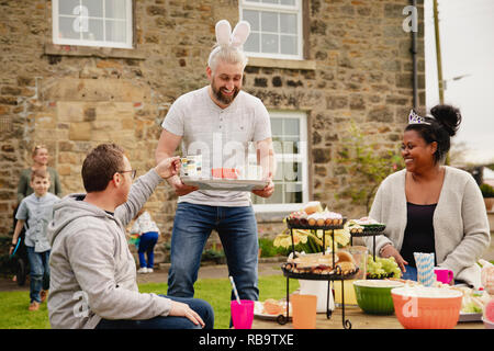 Mid adult man carrying a tray with cups of tea. He is handing out refreshments while wearing rabbit ears at a easter garden party. Stock Photo