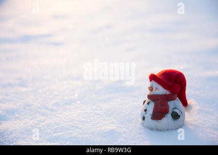 White snowman figure with warm hat and scarf standing in snow, sun setting outdoors in winter, lot of copy space. Stock Photo