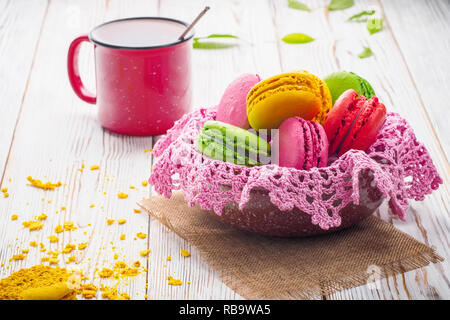 Assorted colorful sweet gentle soft French macaroons dessert cake macarons biscuit in bowl with knitted pink napkin on bright white table with red mug Stock Photo
