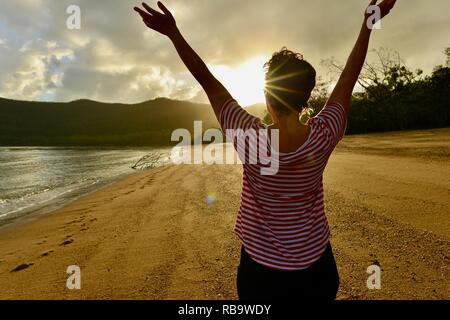Woman holds arms up in freedom to celebrate sunrise on a beach, Smalleys Beach Camp Ground, Cape Hillsborough National Park, Qld, Australia