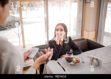 Cheerful young businesswoman sit at table and look at waitress. She smile. Model has bowl of salad on table. Waitress writing order Stock Photo