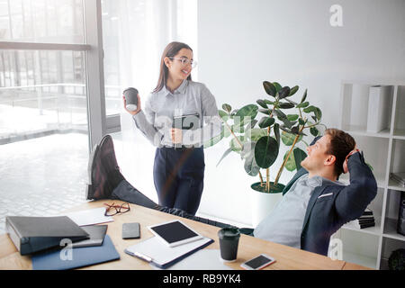 Young woman smiling and flirting with man. She look at him. Model hold cup of coffee and notebook in hands. He sit at table and hold legs there Stock Photo