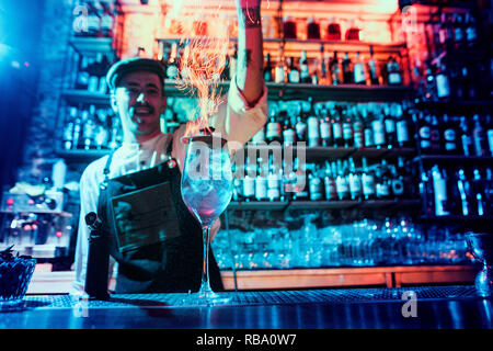 Glass of fiery cocktail on the bar counter against the background of bartenders hands with fire. Barman day concept Stock Photo
