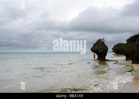 Young boy standing under limestone sculpture by the sea at sandy beach at Ibaruma Sabichi Cave, Ishigaki, Japan
