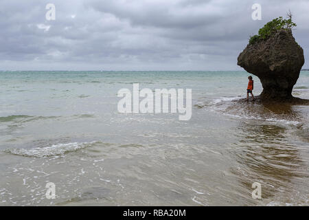 Young boy standing under limestone sculpture by the sea at sandy beach at Ibaruma Sabichi Cave, Ishigaki, Japan