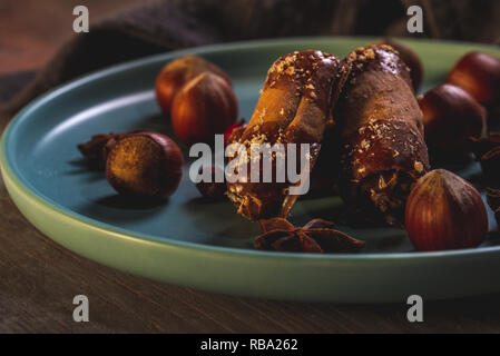 Horizontal photo of few sweet rolls stuffed by cream with chocolate on ends. Sweets are on blue plate with several raw hazelnuts. Plate is on wooden b Stock Photo
