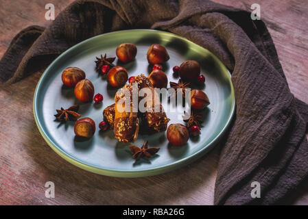 Horizontal photo of few sweet rolls stuffed by cream with chocolate on ends. Sweets are on blue plate with several raw hazelnuts. Plate is on wooden b Stock Photo