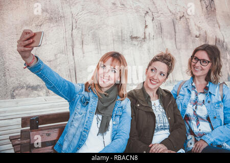 Three happy young women with winter clothing sitting on a wooden bench and taking a selfie Stock Photo