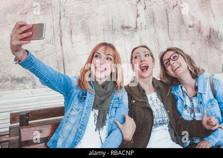 Three happy young women with winter clothing sitting on a wooden bench and taking a selfie Stock Photo