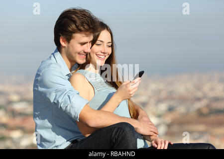 Happy couple checking smart phone content sitting in the city outskirts Stock Photo