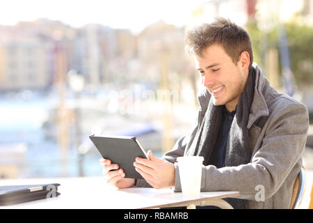 Happy man using a tablet sitting in a coffee shop in a coast town Stock Photo