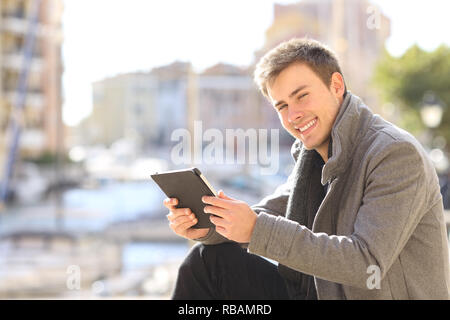 Happy man holds a tablet and looks at camera sitting in the street of a coast town Stock Photo