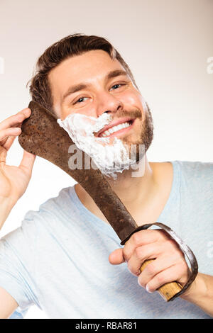 Young man with shaving cream foam on half of face having fun with machete large knife. Handsome guy removing beard hair. Skin care and hygiene humor. Stock Photo