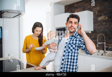 A portrait of young family standing in a kitchen at home, a man making a phone call and a woman feeding a baby. Stock Photo