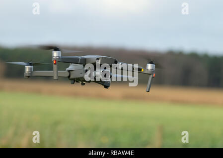 A DJI Mavic 2 Pro quadcopter drone with a mounted camera flies low over a grassy field, with a blurred treeline and cloudy sky in the background Stock Photo