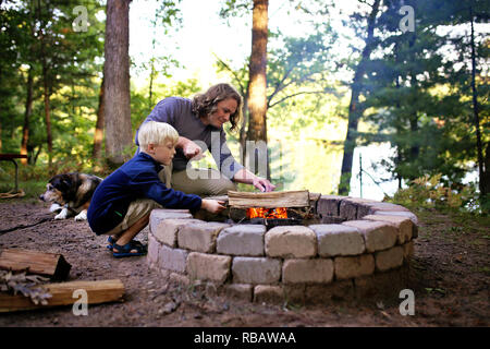 A father and his little son are starting a campfire in a fire ring at a campground overlooking a lake in the woods, as their senior dog lays nearby. Stock Photo