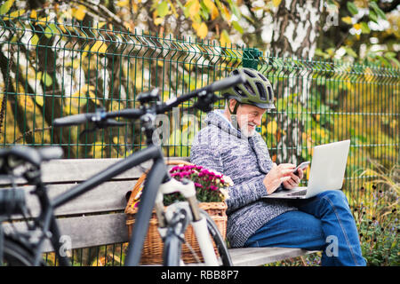 Senior man with electrobike sitting on a bench outdoors in town, using laptop and smartphone. Stock Photo