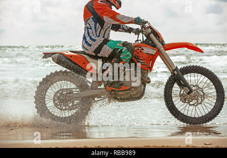 Motorcyclist in a protective suit rides a motorcycle on the sea, splashes fly from under the wheels. Stock Photo