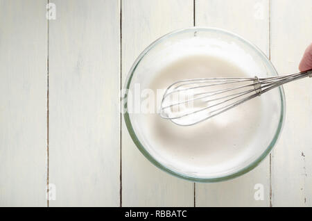 Overhead shot of frothy aquafaba (chickpea water). Vegan egg white replacer in glass bowl with hand holding metal whisk on painted white wood planked  Stock Photo