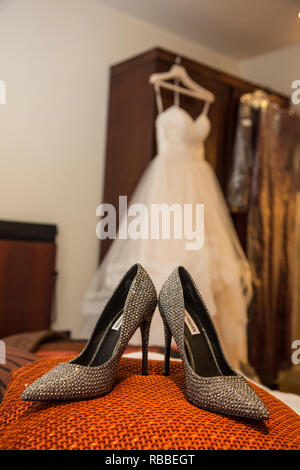 Sparkly Brides shoes on an orange pillow with the brides dress hanging up in the background waiting to be worn on her wedding day Stock Photo