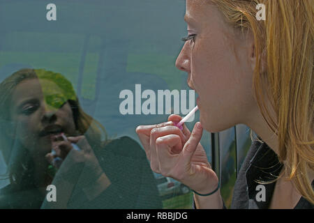 Blonde lady putting on lipstick and using the car window as her mirror Stock Photo